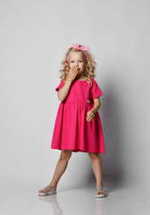 Little blonde kid girl with curly hair in stylish girlish pink dress and headband stands holding hand at mouth, giggling