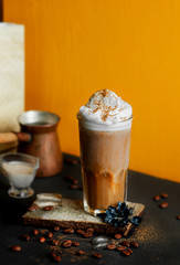 black cold coffee with whipped milk foam and spices in tall glass on natural stone stand, coffee beans, ice cubes, milk jug, Turkish coffee pot on black table and yellow background. Vertically