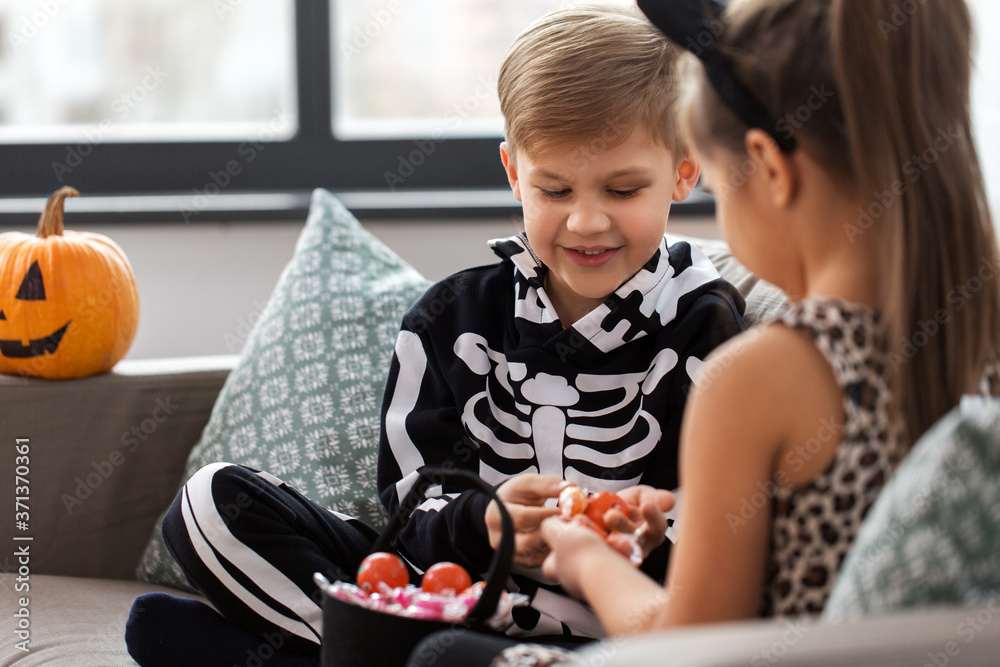 Wall mural halloween, holiday and childhood concept - smiling little boy and girl in party costumes with candies sitting on sofa at home