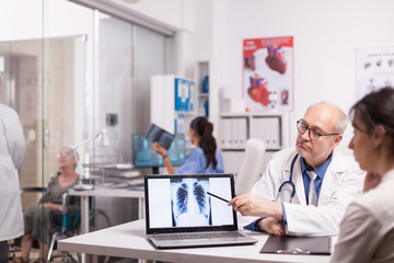 Obraz premium Senior doctor pointing at x-ray of healthy lungs on laptop in hospital office during patient medical visit. Young medic explaining diagnosis to disable mature woman in wheelchair.