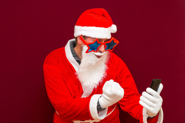 Portrait of satisfied santa claus use cell phone and clenched fistsisolated over red background