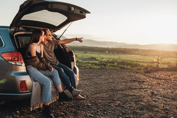 Young Traveler Couple on a Road Trip, Man and Woman Sitting on the Opened Trunk of Their Car Over...