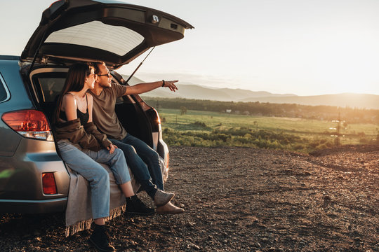 Young Traveler Couple on a Road Trip, Man and Woman Sitting on the Opened Trunk of Their Car Over Sunset