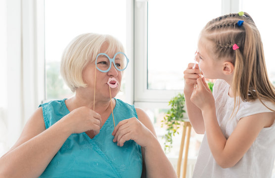 Happy little girl with grandmother putting on glasses and lips on sticks in light room