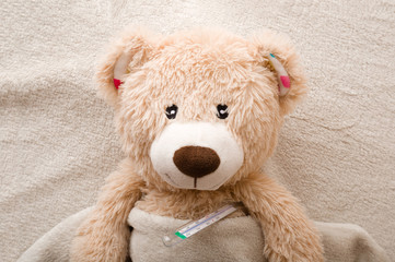 Brown sad teddy bear with mercury thermometer on bed. Children healthcare concept. Closeup. Top...