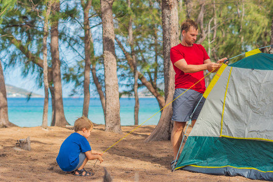 Camping People Outdoor Lifestyle Tourists Putting Up Setting Up Their Green Grey Campsite In Summer Forest Near Lazur Sea. Cute Boy Son Helps His Father Assembling Tent. Natural Education Of Children