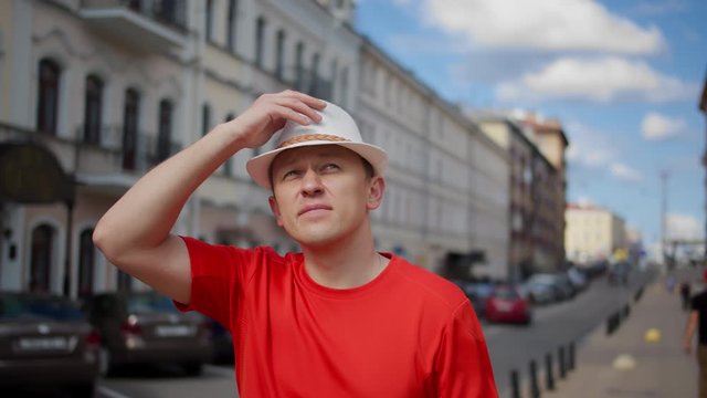 Attractive man in a white hat walks along the city street, examines everything around, tracking with a camera