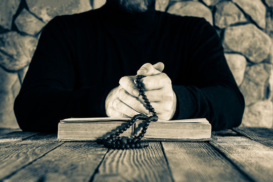 man in black clothes with a rosary in his hands in front of an open old book bible or quran, the concept of prayer, monochrome photo