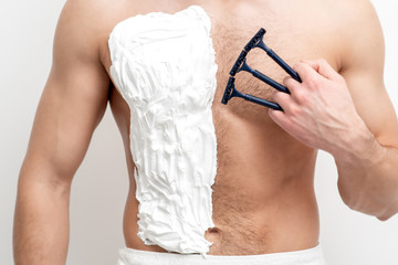 Young caucasian man with beard holds razor shaves his chest with white shaving foam on white background. Man shaving his torso