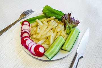 Fried potatoes with spices, chopped radish, cucumbers, green onions, lettuce. All of this lying in a plate standing on a wooden table. Knife and fork lying nearby