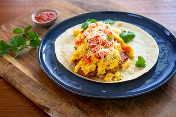 Breakfast burrito with eggs, tomato, bacon and cheese on open tortilla with salsa and coriander or cilantro 