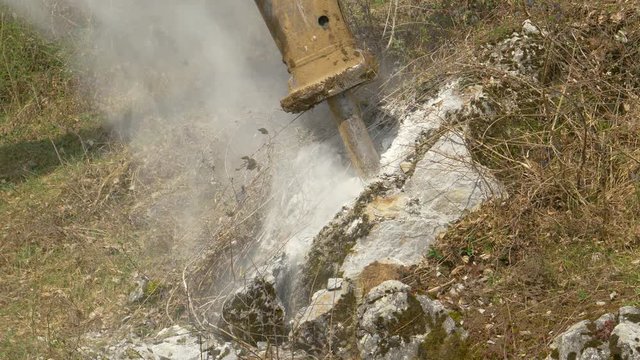 CLOSE UP: Big industrial jackhammer pounds a rock near a meadow at a construction site in the countryside. Dust rises as breaker attachment demolishes rocks during roadworks. Breaker crushing rocks