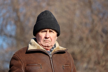 Portrait of elderly man with stern face standing on autumn rural background. Concept of cold...