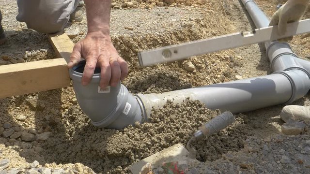 SLOW MOTION, CLOSE UP: Plumbers use a level and fresh concrete to bury a wide grey sewage pipe. Unrecognizable contractors assemble and bury a sewage system for a nearby house under construction.