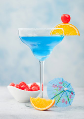 Blue lagoon summer cocktail in margarita glass with sweet cocktail cherries and orange slice with umbrella on blue background.
