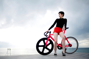 Full length portrait of professional female cyclist rider dressed in red sport shorts enjoying cloudy sky calm scenery while leaning on her fixed gear bicycle against copy space cloudy background
