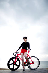 Obraz premium Portrait of professional female cyclist enjoying the rest after bike ride along seashore while standing on the pier against sea and sky background with copy space area for your text message or content