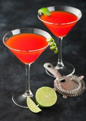 Cosmopolitan cocktail in classic crystal glasses with lime peel and fresh limes with strainer on black background.