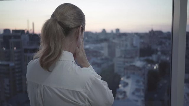 Depressed mid-adult businesswoman standing in front of window looking at city in the evening. Back view of stressed Caucasian woman standing on balcony and thinking at dusk. Overworking concept.