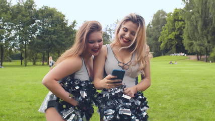 Two cheerleading girls having fun surfing internet on smartphone and laughing standing outdoors