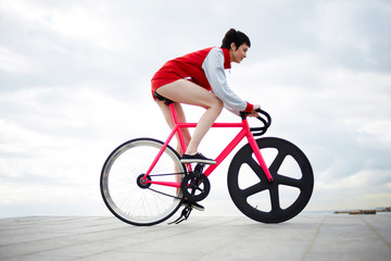 Portrait of a young sports woman dressed in stylish red tracksuit riding on light weight fixed gear bicycle on concrete pier seashore against sky copy space area for you text message or information