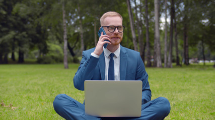 Young businessman with laptop talking on smartphone sitting on green grass in park