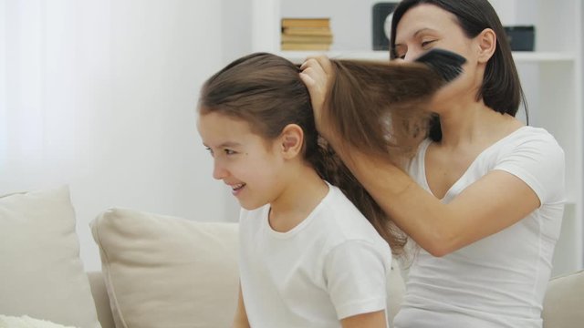 The mother is combing her daughter, doing her a ponytail hairdo.