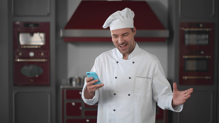 Happy chef standing in modern kitchen reading exciting feedback from client on smartphone