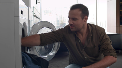 Young Man Loading Clothes Into Washing Machine In Kitchen