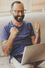 Excited young business man student doing winner gesture, sitting with laptop on sofa at home,closed...