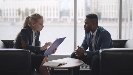 Young HR woman interviews afro-american male candidate for job sitting on couch in conference room