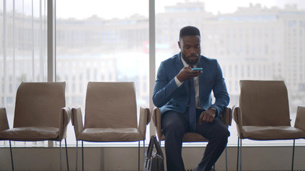 Afro entrepreneur wait for meeting with partners sitting on chair sending voice mail on smartphone