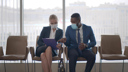 Colleagues in protective mask discussing project plan on clipboard sitting in hall