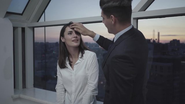 Confident businessman touching hair of beautiful brunette colleague standing on glazed balcony in the evening. Portrait of Caucasian man and woman flirting in office at dusk. Love affair concept.