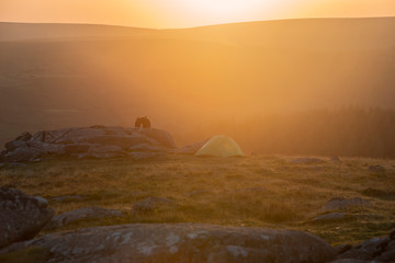 Stunning image of unidentified young couple wild camping in English countryside watching the stunning Summer sunrise with warm glow of the sun lighting the landscape