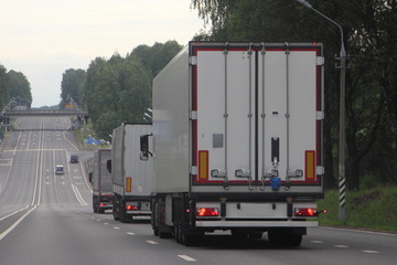 A many white vansemi trucks lorry convoy drive next to 2-lane asphalted country road on forest background on summer day, back view, transportation Logistics, international commercial freight by road