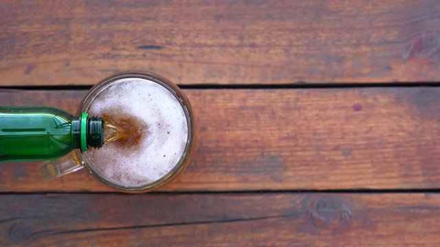 Closeup top view flatlay 4k video of pouring fresh beer from big plastic green bottle into big glass mug standing on brown rustic wooden table background.