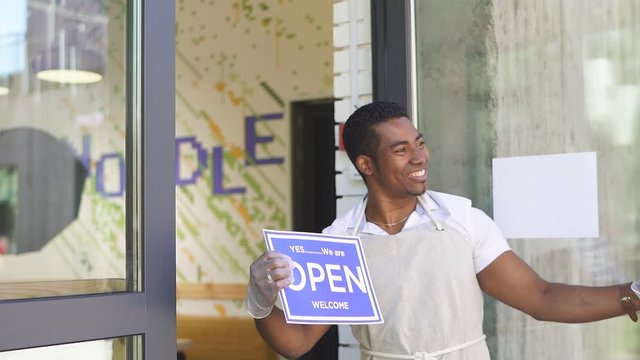 You are welcome. cafe worker welcomes clients, open-minded black man in uniform smile, invites customers in shop.