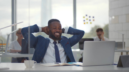 Happy afro businessman with hands behind head smiling resting at desk in office.