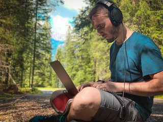 young brunette caucasian man with beard wearing headphones and working on laptop in a mountain place, green forest in the background