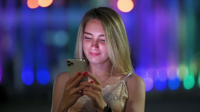 Cute Girl Uses Smartphone and Smiles Standing on Night Street in the City. Woman Messaging on Phone at Night on the Street With the Lights of the Road Blurred Background.