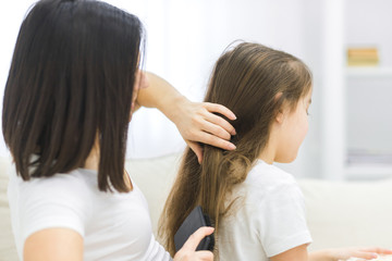 Close-up photo of mother making hair style to her daughter in light white interior room.