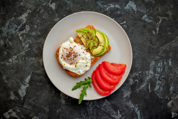 Toast with poached egg, flaxseeds, avocado and tomato slices, arugula leaves on a plate. Healthy meal for breakfast.
