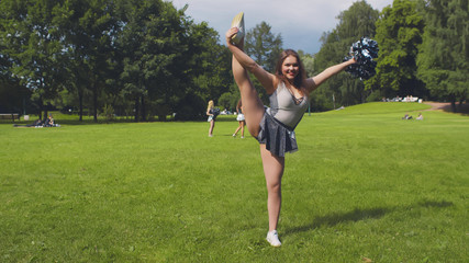 Beautiful cheerleader in uniform practicing split holding pompom standing outdoors