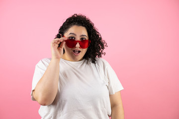 Young beautiful woman wearing sunglasses over isolated pink background surprised and taking and lowering her sunglasses