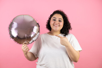 Young beautiful woman standing over isolated pink background smiling. Pointing and holding a balloon