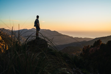 A man stands on a rock looking to the right while trekking during golden light on a mountain looking at the stunning views of Serra de Tramuntana (Mallorca, Spain)