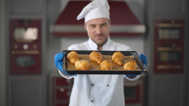 Handsome baker in uniform holding tray full of freshly baked croissants at modern kitchen