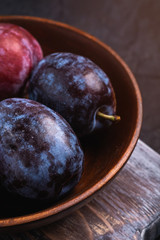 Fresh sweet plum fruits in brown wooden bowl on old cutting board, black textured background, angle view macro