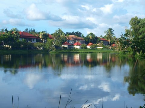 Beautiful blue sky with white cumulus clouds reflected on the water surface. Houses on the shore. Greenery, pond, view of the village on the shore. Countryside panorama. Trees, grass, palm, cottages. 